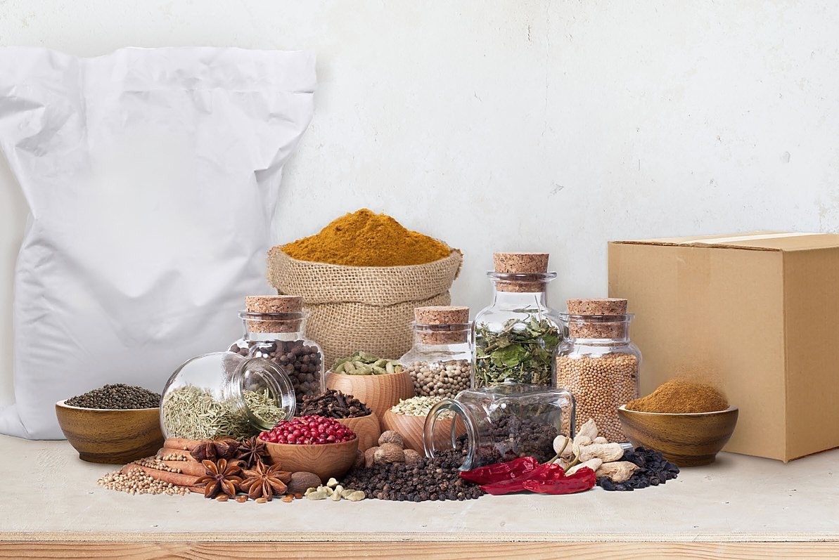 Whole and ground spices in jars and wooden bowls in front of a bulk spice sack and a carton